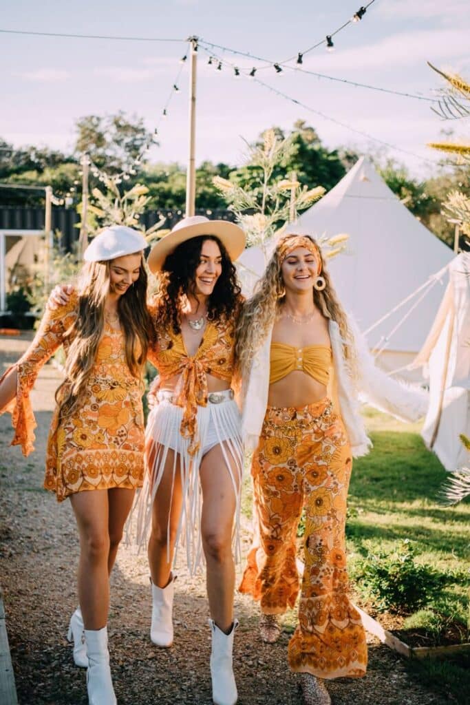 Three women in bohemian outfits enjoying an outdoor festival with tents in the background.