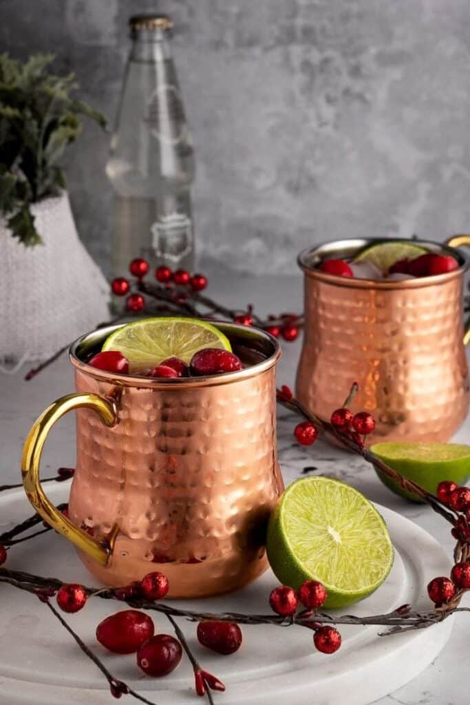 Copper mugs with lime and cranberries on a tray, bottle in the background; festive, rustic drink setting.