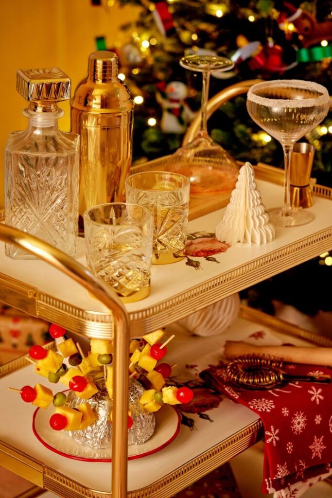 Festive holiday bar cart with crystal decanter, gold shaker, and snacks, set against a decorated Christmas tree background.