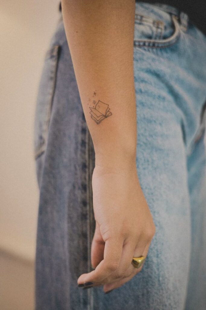 Fine line magic book tattoo Minimalist book tattoo on forearm, wearing jeans and a gold ring. | Sky Rye Design Minimalist book tattoo on forearm, wearing jeans and a gold ring.