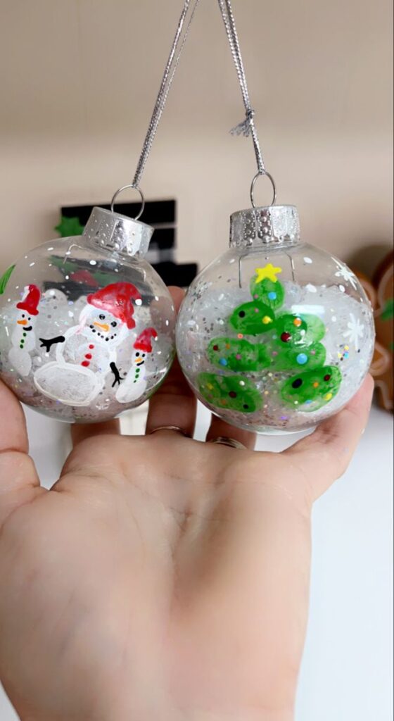 Hand holding Christmas ornaments: one with snowmen, the other with a decorated tree. Festive holiday decor.