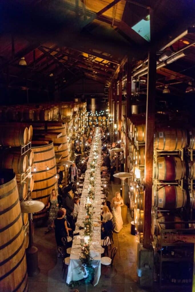 Wedding reception in a winery with long table adorned with lights, surrounded by barrels. Romantic and elegant setting.