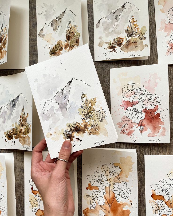 Hand holding watercolor artwork of mountains and flowers, surrounded by similar nature-themed paintings on a wooden surface.