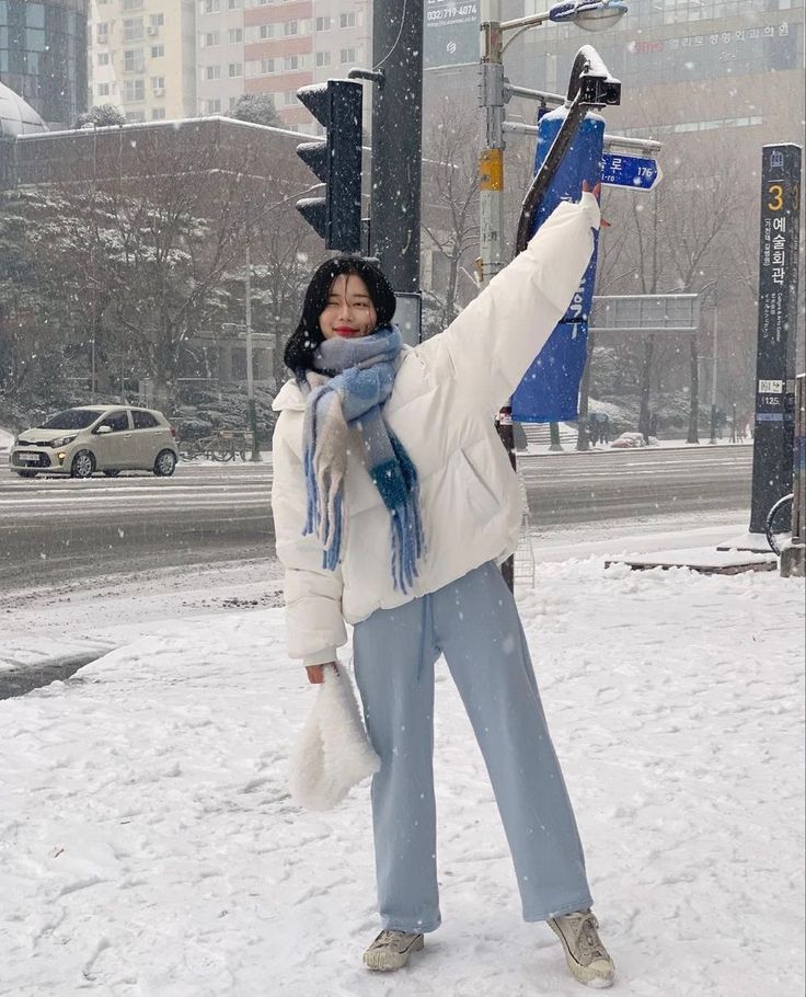Person in winter attire posing joyfully in snowy city street with buildings in background.