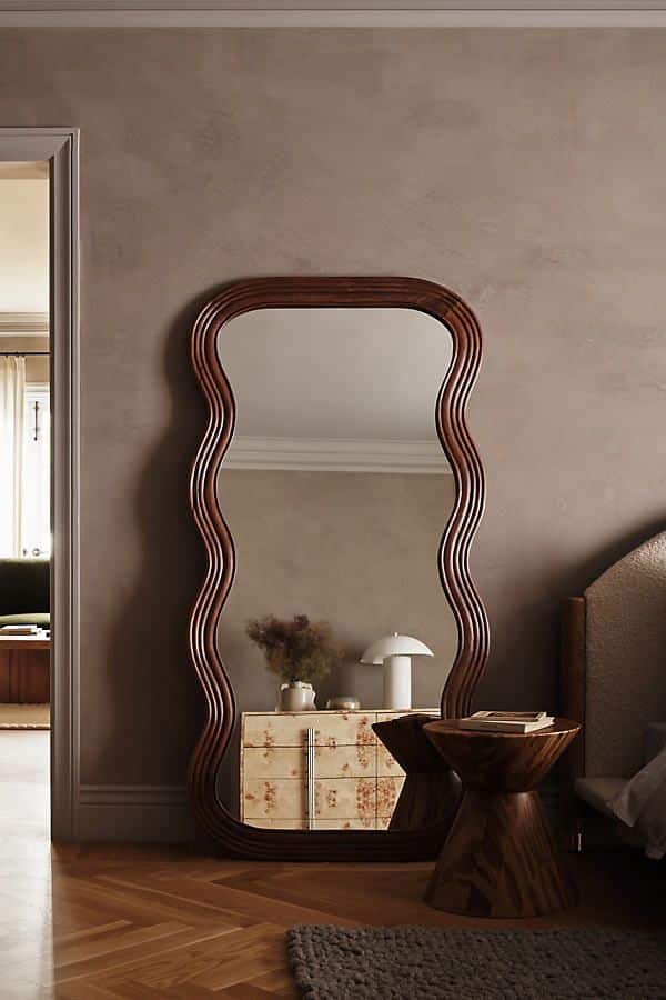 Wavy wooden-framed mirror in cozy bedroom with chic decor, side table, and beige walls. Modern interior design.