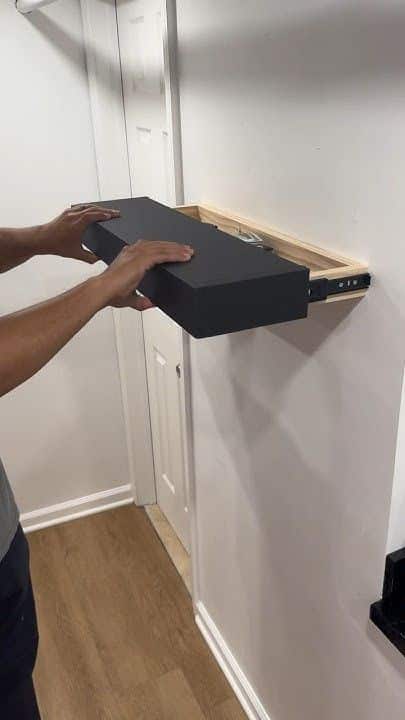 Person installing a black floating shelf on a white wall with wooden supports.