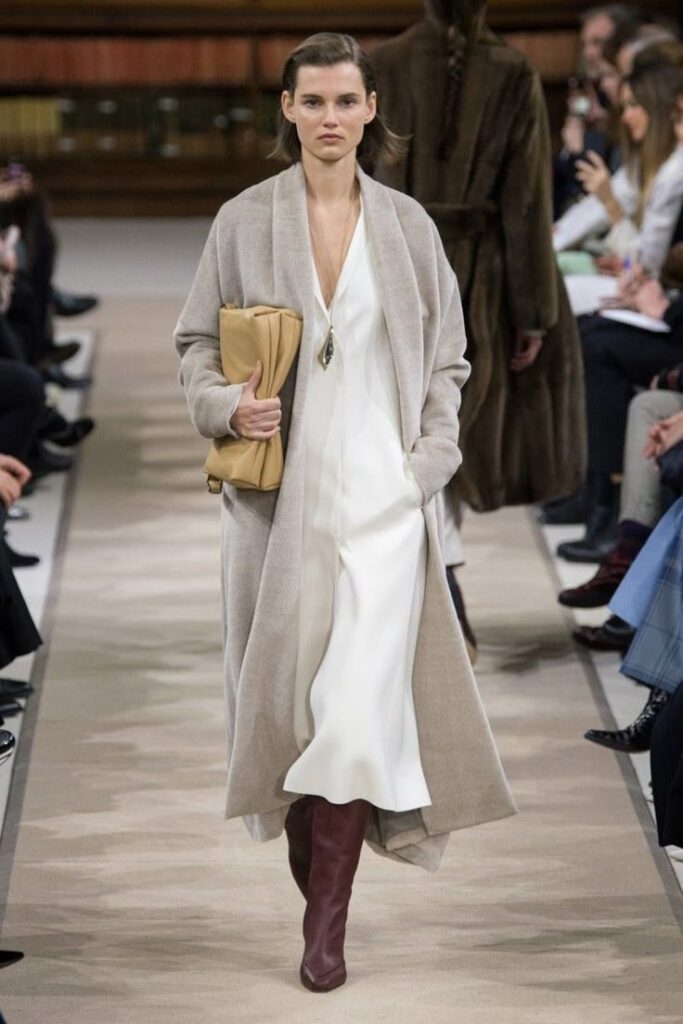 Model walking runway in beige coat, white dress, and brown boots at fashion show.