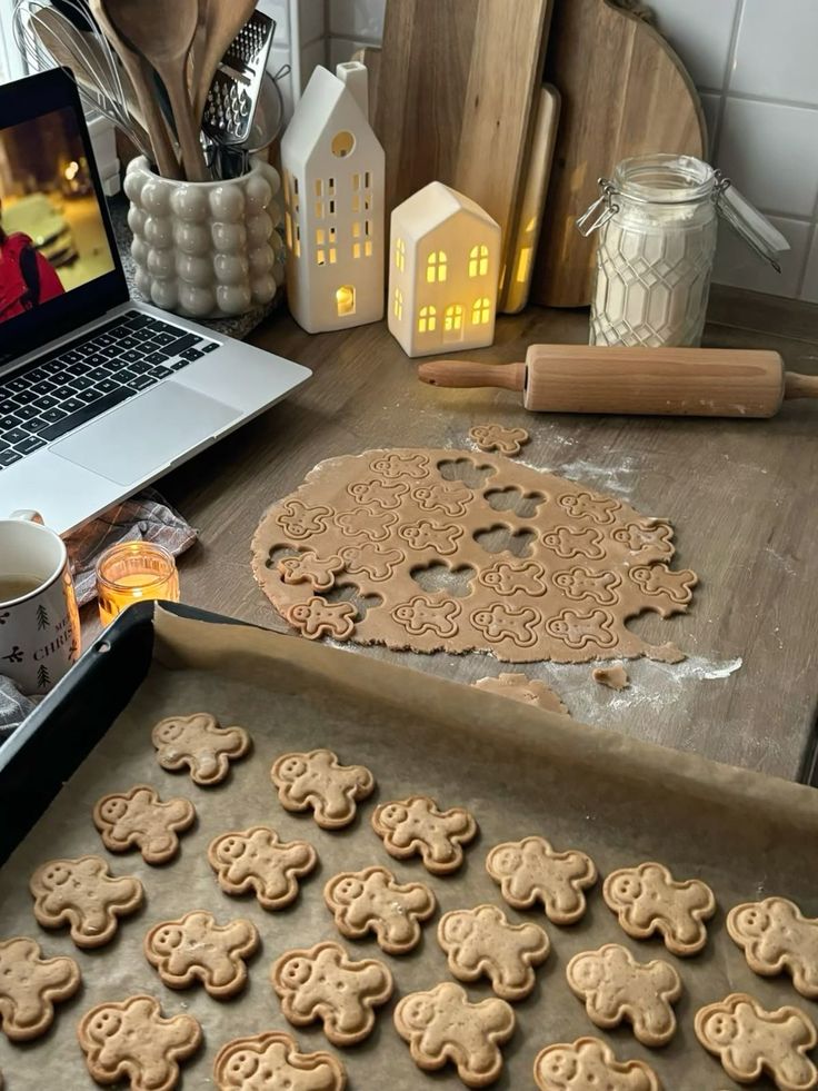 Cozy kitchen scene with gingerbread cookies being shaped and baked, surrounded by warm lights and a laptop.