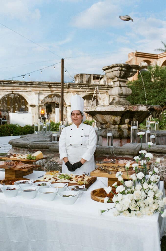 Chef in uniform stands by elegant outdoor buffet table with assorted dishes and flowers. Stone fountain in background.