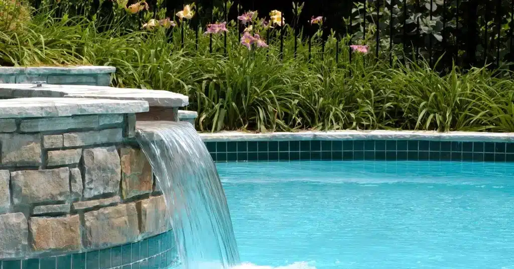 GulfCoastPoolConsultingConstruction-370447-Pool-Waterfall-Flowers-image-a1jpeg Stone waterfall feature cascading into a serene blue pool, surrounded by lush greenery and blooming flowers. | Sky Rye Design Stone waterfall feature cascading into a serene blue pool, surrounded by lush greenery and blooming flowers.