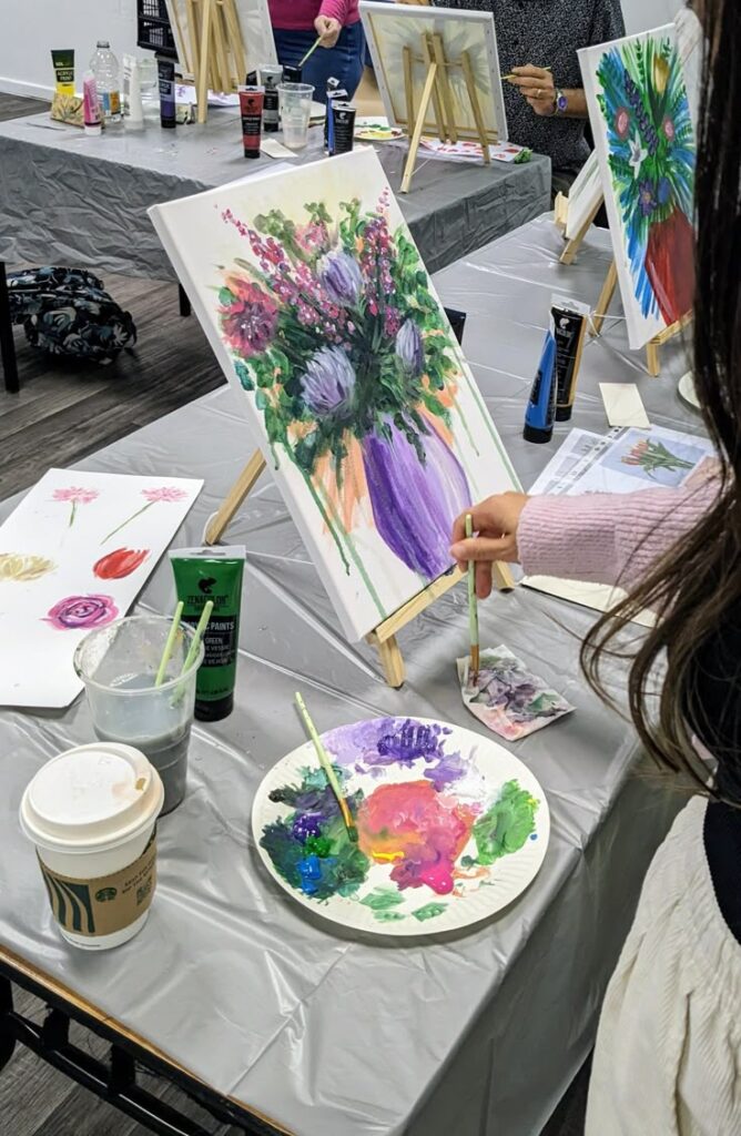 HOST YOUR OWN PAINTING WORKSHOP - Any theme - First 6 guests Person painting colorful flowers on canvas at a paint class, with brushes and paints on the table. | Sky Rye Design Person painting colorful flowers on canvas at a paint class, with brushes and paints on the table.