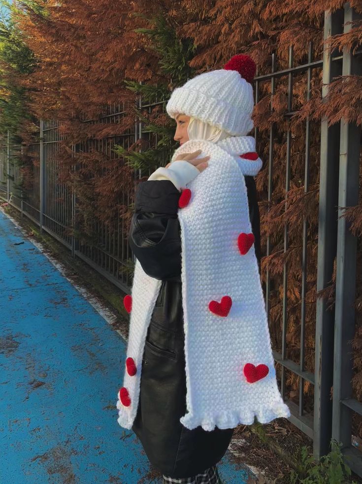 Handmade Knit Scarf Beanie Set White with Red 3D Hearts Person in winter attire with a white knit scarf and beanie, adorned with red hearts, walking along a scenic pathway. | Sky Rye Design Person in winter attire with a white knit scarf and beanie, adorned with red hearts, walking along a scenic pathway.