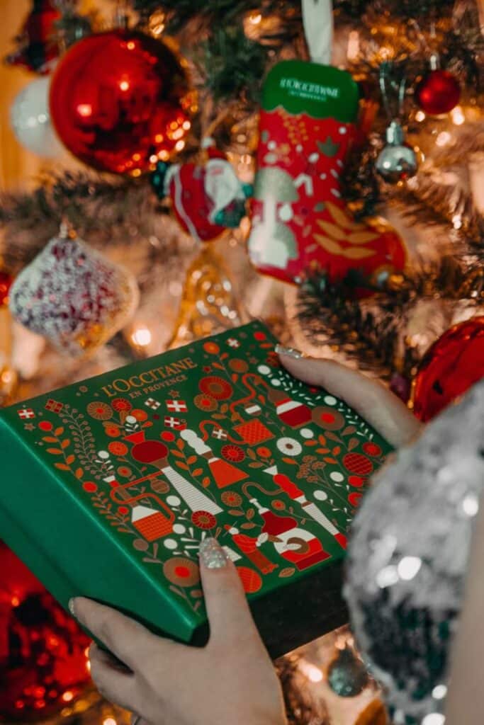 Happy New Year 🎄✨ Hands holding a festive gift box in front of a decorated Christmas tree with ornaments and lights. | Sky Rye Design Hands holding a festive gift box in front of a decorated Christmas tree with ornaments and lights.