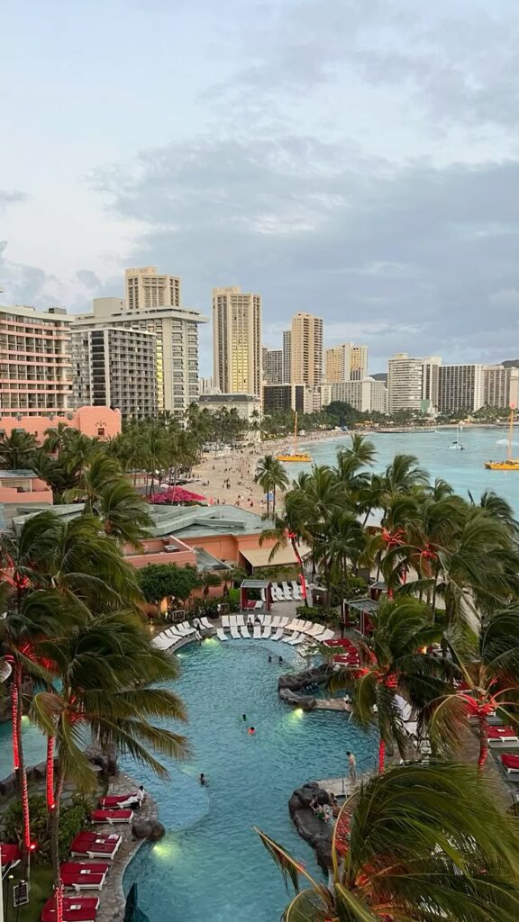 Aerial view of tropical resort with pool, palm trees, and city skyline by the ocean.