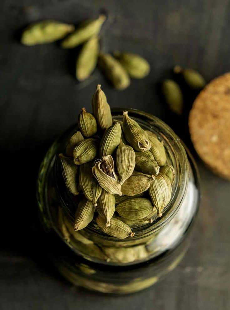 Hel Spice - Iranian Green Cardamom + 6 Benefits Glass jar filled with green cardamom pods on a dark background, with some pods scattered around. | Sky Rye Design Glass jar filled with green cardamom pods on a dark background, with some pods scattered around.