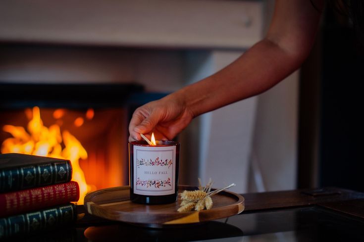 Hello Fall _ Autumn Scented Candles _ Cosy Candle _ Aromatherapy _ Gifts _ Housewarming _ Clean Burning _ Autimn Decor _ Home Decor _ Hand lighting a cozy fall candle on a wooden tray by the fireplace, with books and dried flowers nearby. | Sky Rye Design Hand lighting a cozy fall candle on a wooden tray by the fireplace, with books and dried flowers nearby.