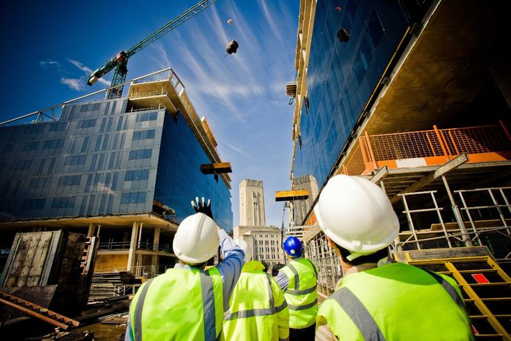Construction workers in safety gear observe crane operation at urban building site under a clear blue sky.