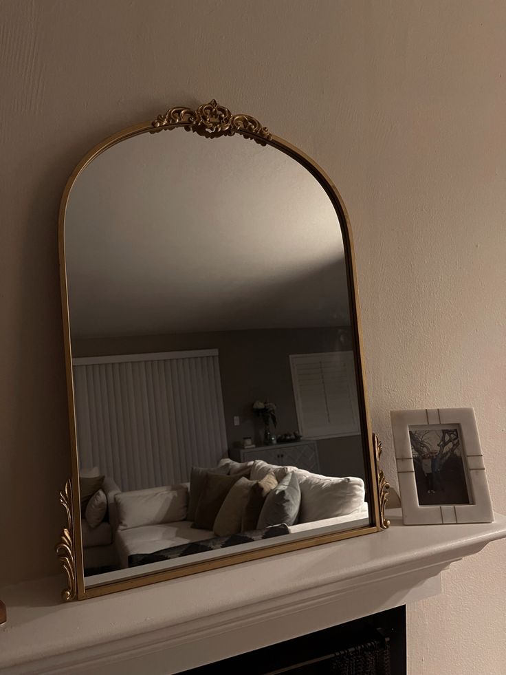Elegant gold-framed mirror on mantel, reflecting cozy living room with soft pillows and closed blinds.