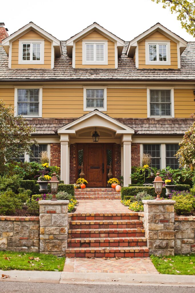 Homeexterior-2CQQ4556KpeBO1h9aQTRT0-8cb7bda414794cbf8a8373ecf4a20a25 Charming yellow house with wood shingles, autumn decor, and landscaped entrance in a cozy neighborhood. | Sky Rye Design Charming yellow house with wood shingles, autumn decor, and landscaped entrance in a cozy neighborhood.