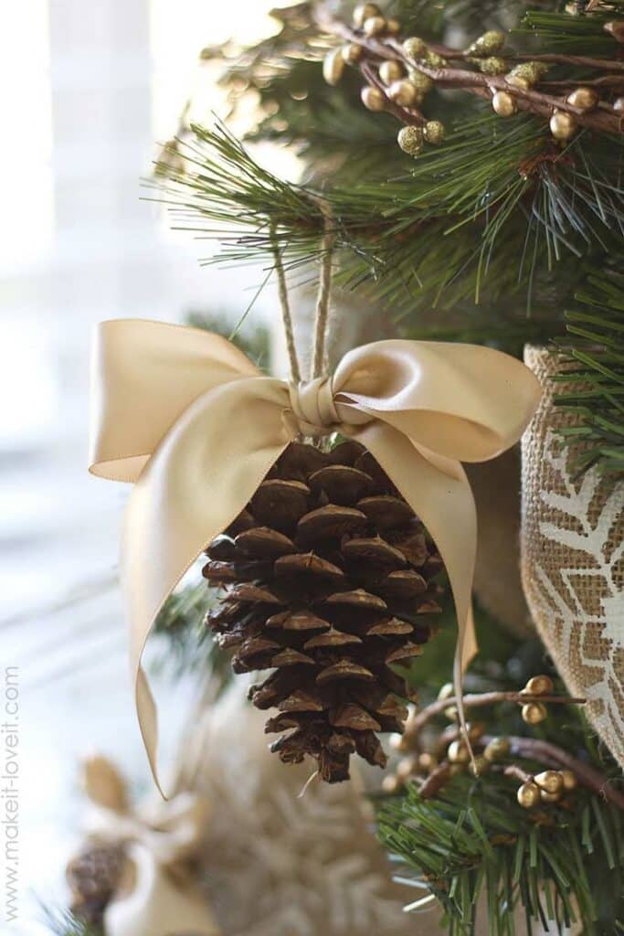 Pine cone Christmas ornament with a gold ribbon bow, hanging on a green tree branch, festive holiday decor.