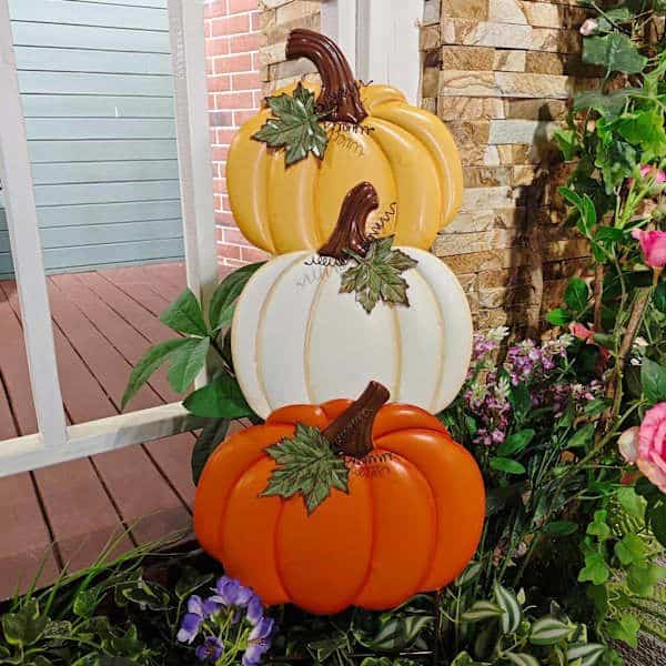 Stacked decorative pumpkins with autumn colors, surrounded by vibrant flowers, next to a porch.