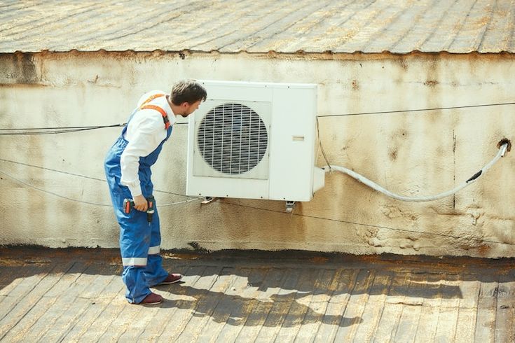 Technician inspecting rooftop air conditioning unit, wearing blue overalls and holding a tool on a sunny day.