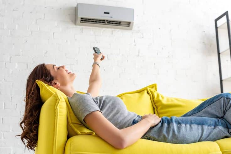 Woman relaxing on a yellow sofa, adjusting air conditioner with remote, white brick wall background.