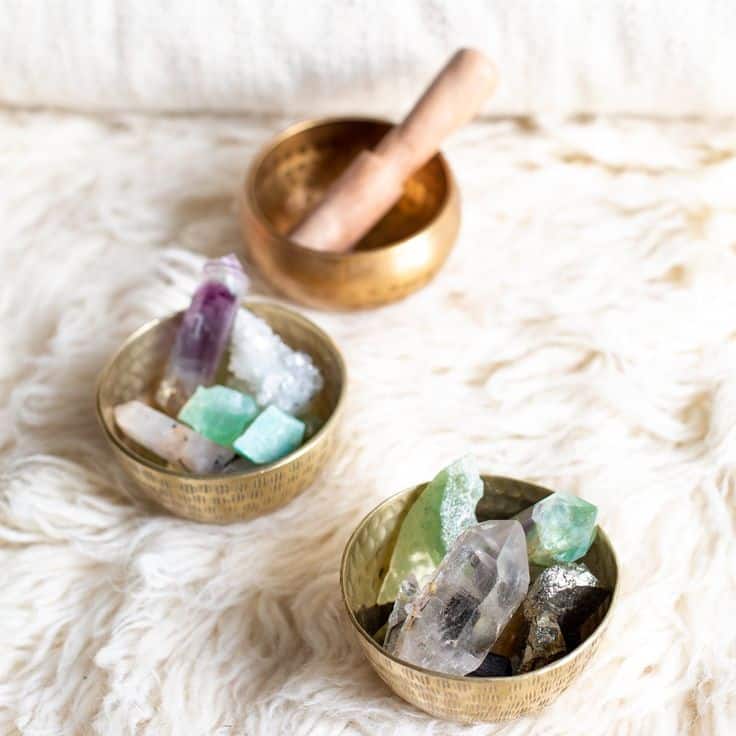 Brass bowls with colorful healing crystals and a singing bowl on a soft, textured fabric surface.
