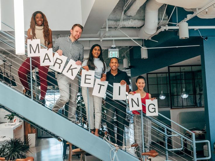 How to Use ChatGPT for Marketing_ Boost Your_ Diverse team on stairs holding MARKETING signs in a modern office setting, symbolizing collaboration and innovation. | Sky Rye Design Diverse team on stairs holding MARKETING signs in a modern office setting, symbolizing collaboration and innovation.