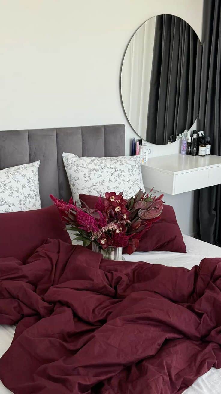 IG_ vlsva _ TG_ vlsvmood Cozy bedroom with burgundy bedding, floral pillows, and decorative vase, accented by a round mirror and stylish decor. | Sky Rye Design Cozy bedroom with burgundy bedding, floral pillows, and decorative vase, accented by a round mirror and stylish decor.