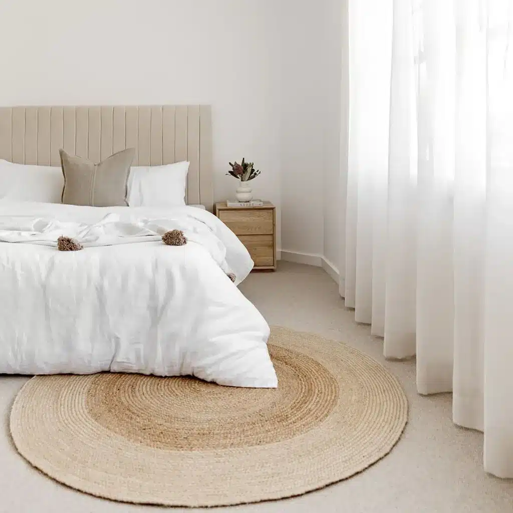 | Sky Rye Design Modern bedroom with beige upholstered bed, white linens, round jute rug, and natural light from sheer curtains.