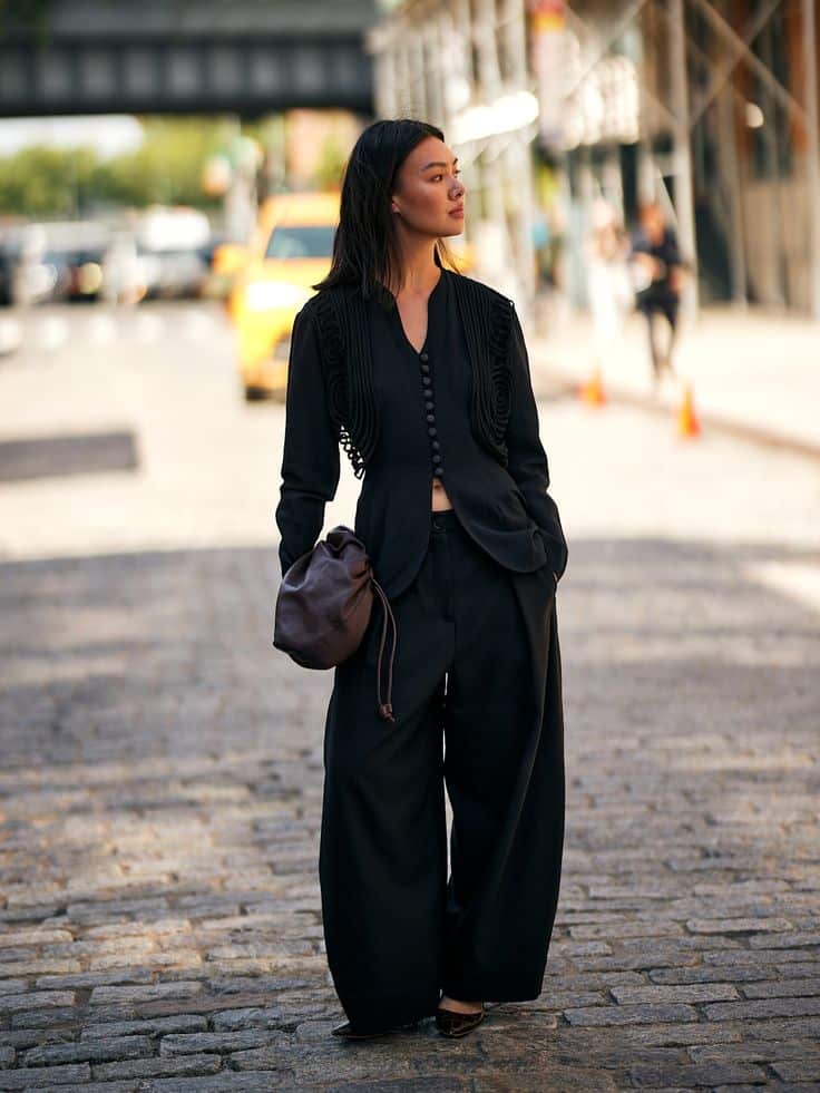 Woman in a stylish black outfit walking confidently on a city cobblestone street, carrying a handbag.