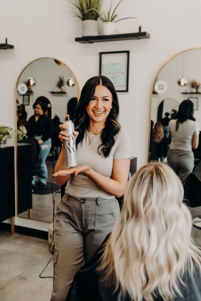 Hairstylist holding hair spray in salon with client, smiling and ready to style hair.