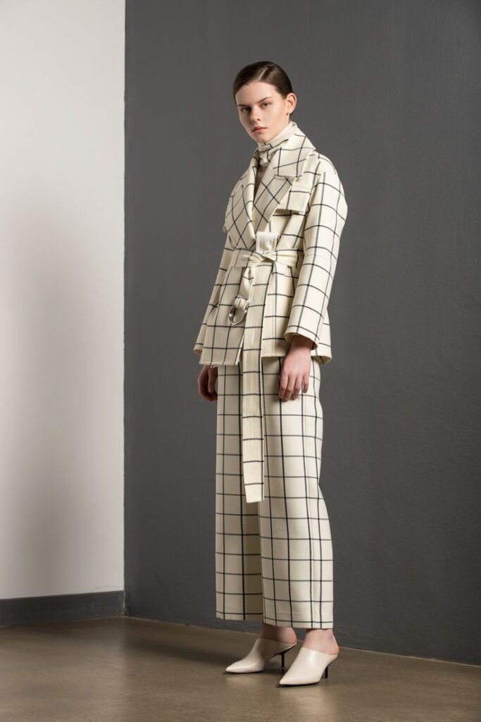 Model wearing a stylish cream and black checkered suit with wide-leg pants and high heels, posed against a gray wall.