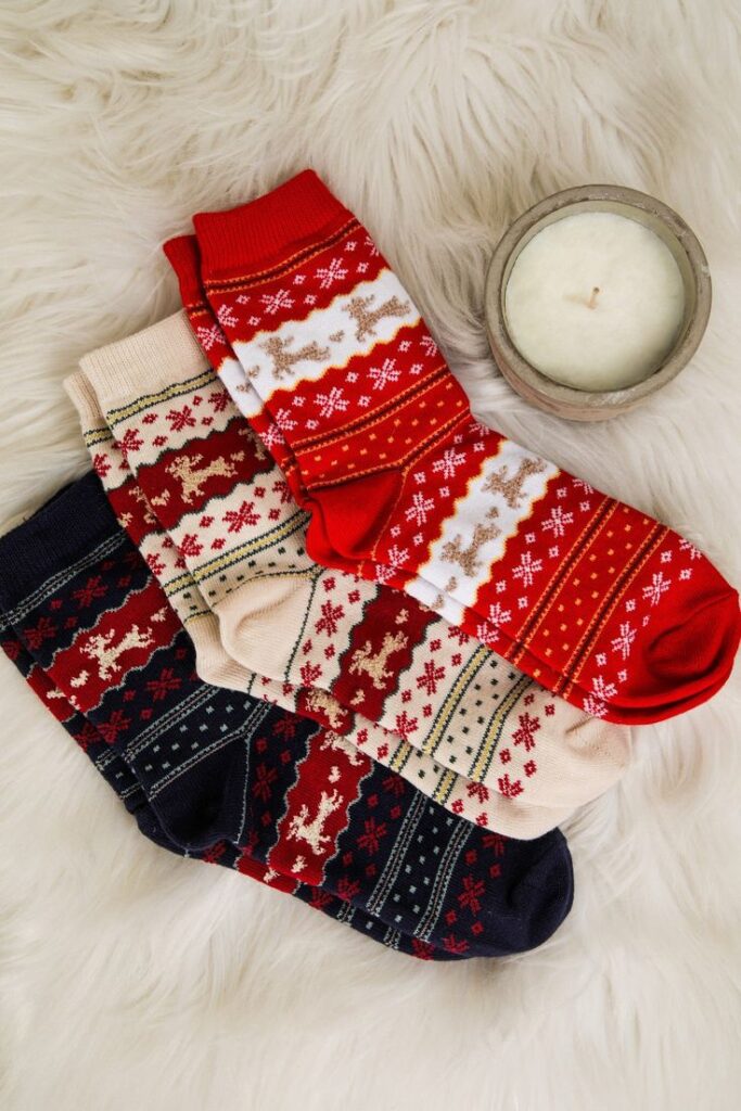Jolly n Bright Socks - Cream Festive socks with reindeer and snowflake patterns on fluffy surface beside a lit candle, cozy winter vibe. | Sky Rye Design Festive socks with reindeer and snowflake patterns on fluffy surface beside a lit candle, cozy winter vibe.