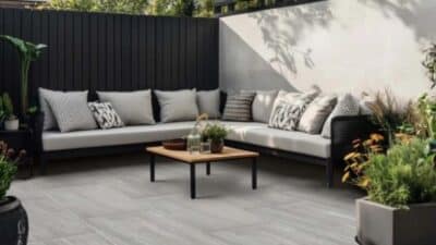 Modern outdoor patio with gray sofa, cushions, and wooden table, surrounded by plants and a white privacy wall.