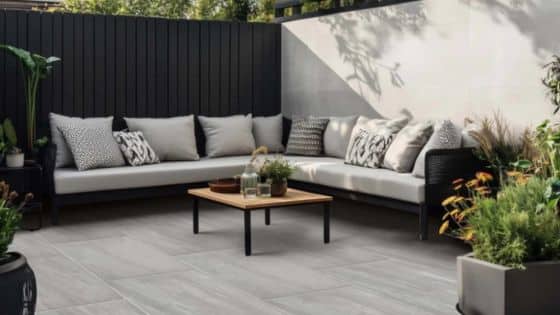 Kandla Grey Porcelain Best Finish for Outdoor Space Modern outdoor patio with gray sofa, cushions, and wooden table, surrounded by plants and a white privacy wall. | Sky Rye Design Modern outdoor patio with gray sofa, cushions, and wooden table, surrounded by plants and a white privacy wall.