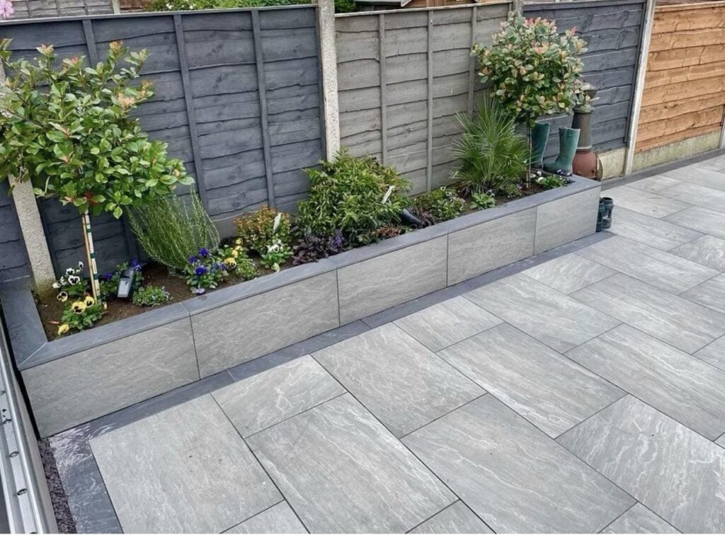 Modern garden patio with gray tiles and lush green plants in a raised flowerbed against a wooden fence.
