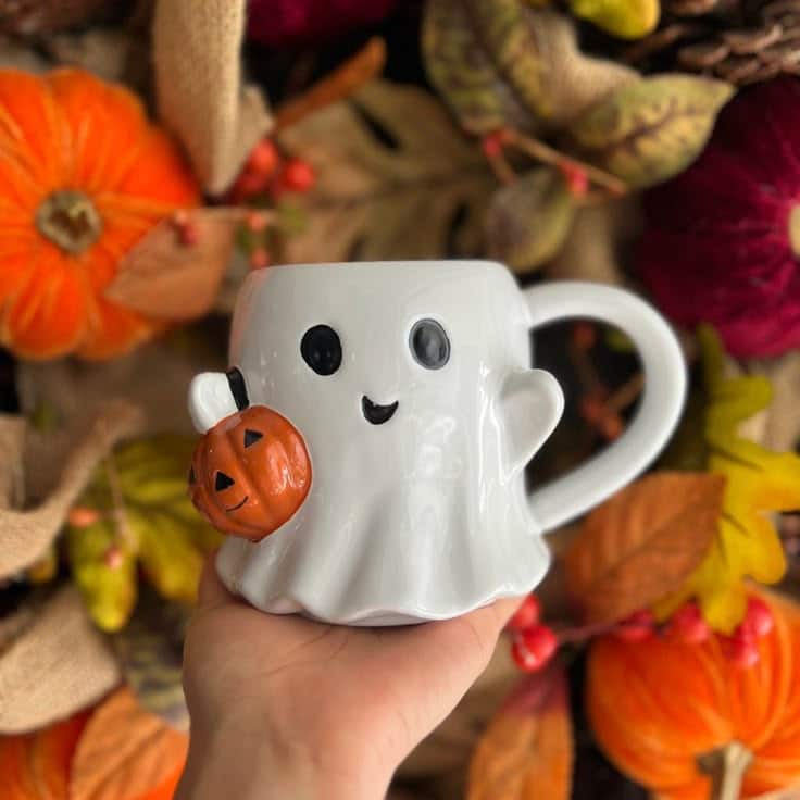 Cute ghost mug with pumpkin design, perfect for Halloween. Surrounded by autumn leaves and festive decor.