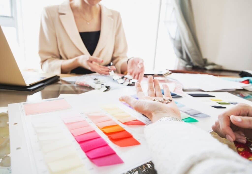Konzultace-s-osobnim-stylistou Fashion designers reviewing colorful fabric swatches and beads in a creative workspace. | Sky Rye Design Fashion designers reviewing colorful fabric swatches and beads in a creative workspace.