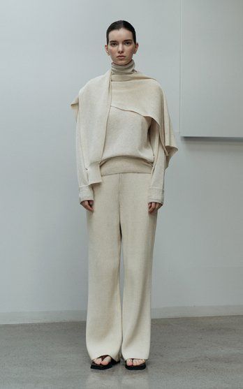 Model in beige turtleneck and wide-leg pants ensemble, standing against a minimalist background.