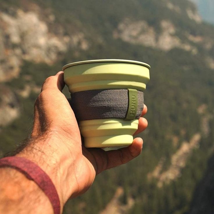 Hand holding a collapsible green camping cup with scenic mountain view in the background.