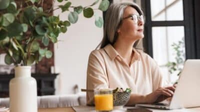 Woman with gray hair using a laptop, seated at a table with a salad and orange juice, near a window with plants.