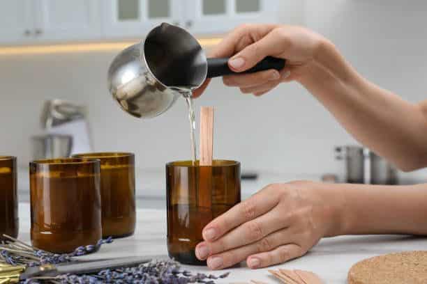 Woman making homemade candle at table in kitchen closeup Pouring wax into brown glass jar with wooden wick for homemade candle making on a white kitchen counter. | Sky Rye Design Pouring wax into brown glass jar with wooden wick for homemade candle making on a white kitchen counter.