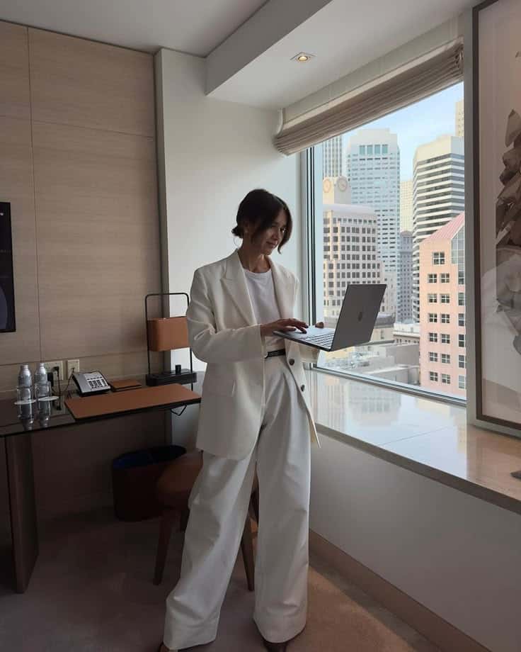 Businesswoman in white suit working on a laptop by a window with city skyline.