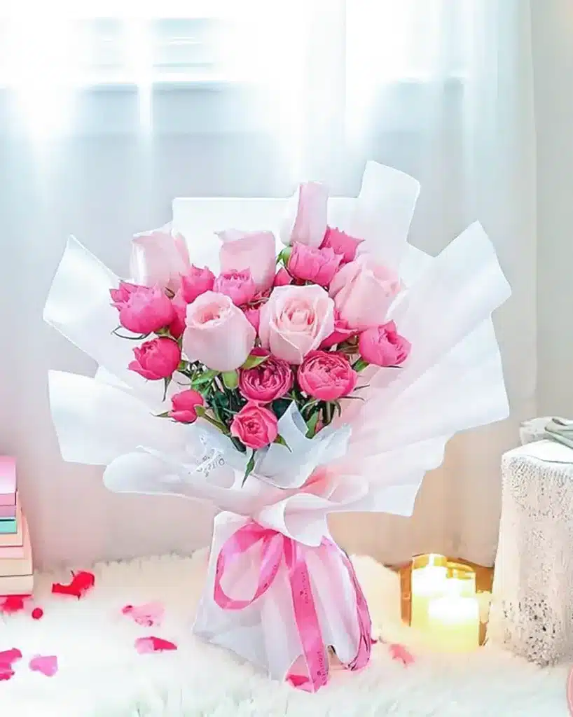 Bouquet of pink roses wrapped in white paper with pink ribbon, set in a cozy room with candles and books.