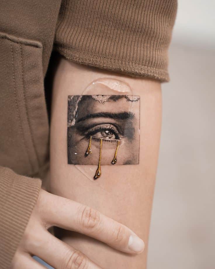 Tattoo of a detailed eye with gold dripping tears on forearm, against a brown sleeve backdrop.