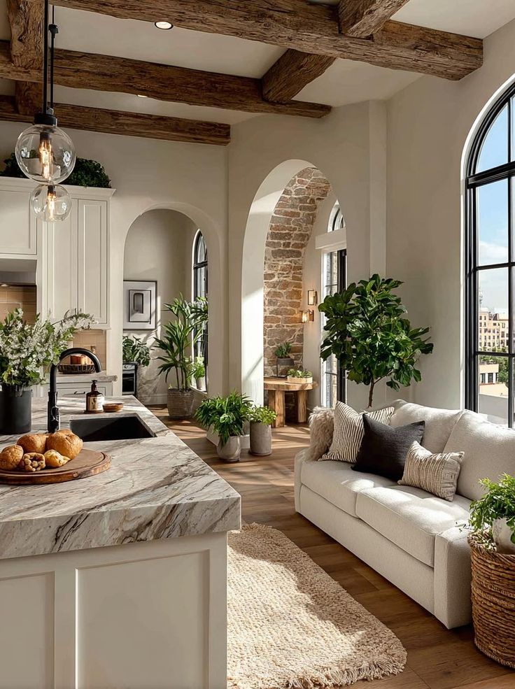 Bright rustic kitchen with wooden beams, marble island, cozy sofa, and lush green plants by arch windows.