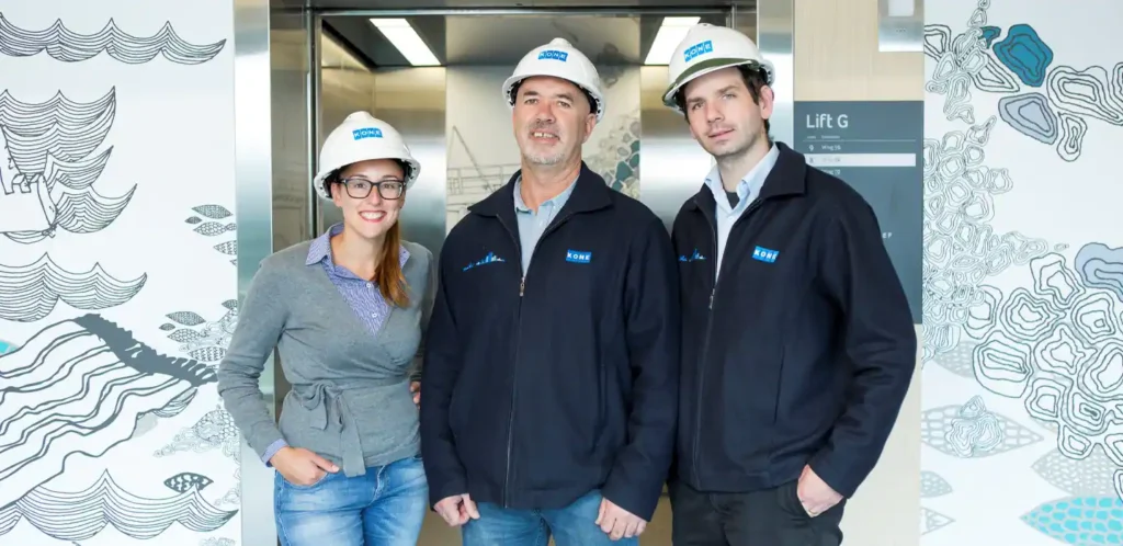 Making_a_better_hospital_hero_1440x700_tcm17-60875 Three engineers wearing Kone uniforms and hard hats smiling in front of an elevator. | Sky Rye Design Three engineers wearing Kone uniforms and hard hats smiling in front of an elevator.