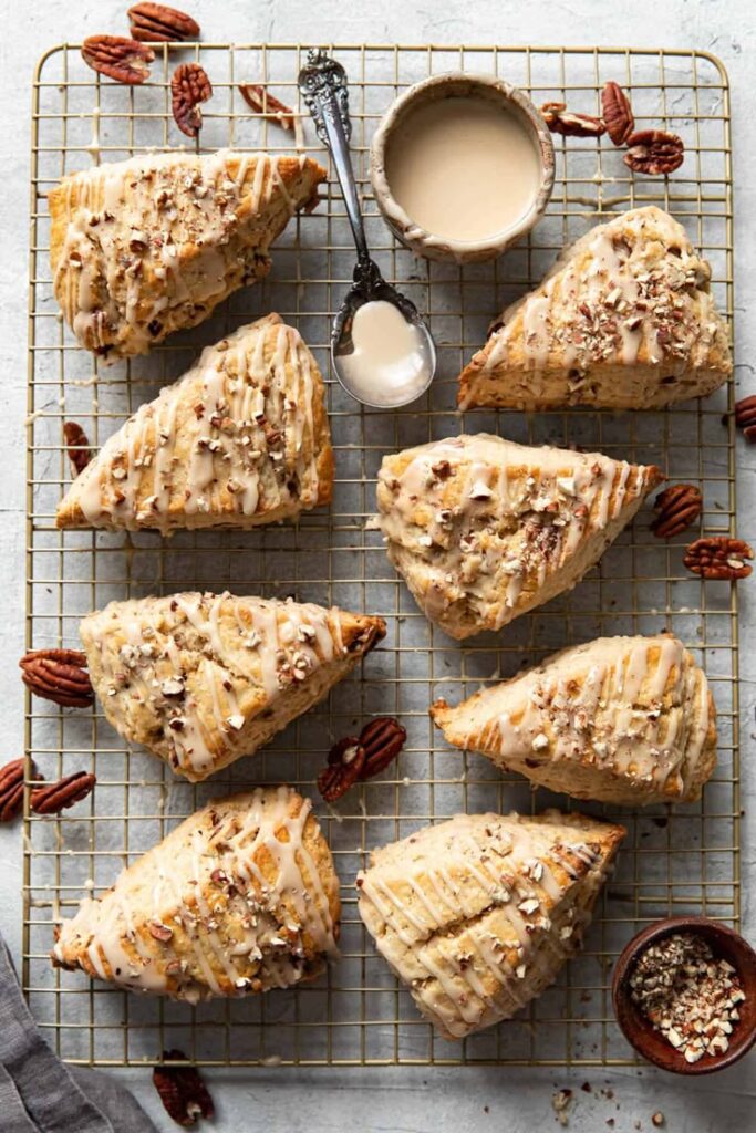 Maple Pecan Scones Pecan scones drizzled with glaze on a cooling rack, surrounded by pecans, ready to serve with extra glaze and chopped nuts. | Sky Rye Design Pecan scones drizzled with glaze on a cooling rack, surrounded by pecans, ready to serve with extra glaze and chopped nuts.